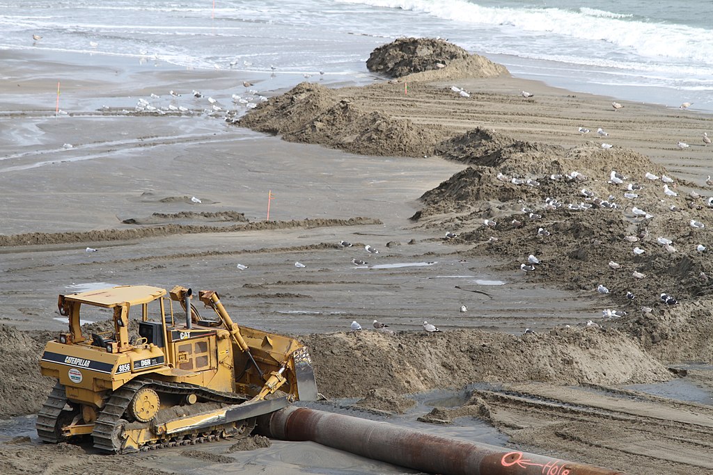 Part of the Virginia Beach Hurricane Protection Beach Re-nourishment Project, a hopper dredges sits off the Virginia Beach oceanfront pumping sand from the ocean floor to a pump-out landing station where the material is piped onshore (by Pamela Spaugy, Army Corps of Engineers CC BY 2.0 via Flickr).
