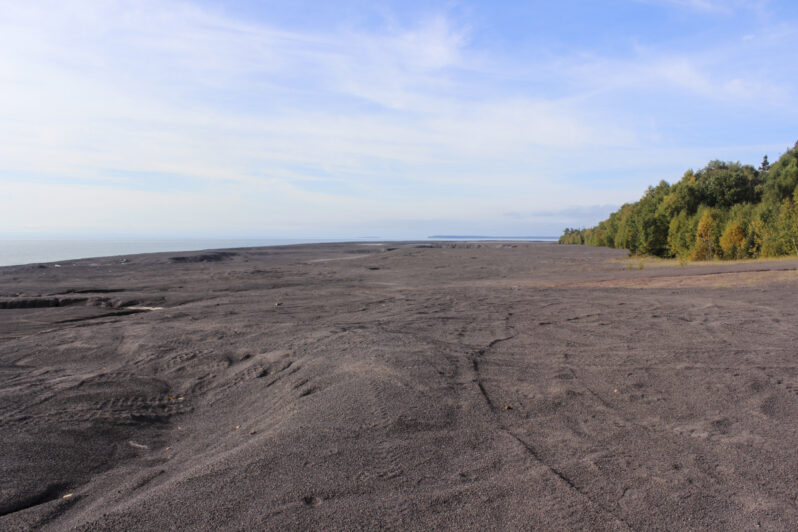 Figure 12. The stamp sand beach in area of spit-widening is typical of the entire length of the landform from Gay to the Traverse River jetties – barren, lifeless, marked by an irregular surface of the remnant spit topography, some man-made excavation pits, and the tracks of the last visit by ORVs. Note that the original accumulation began at about the present tree line although initial over-wash of stamp sand into the forest probably altered plant growth. On a natural Great Lakes beach with this extent of exposure there would have been plant colonization by pioneer plants and a progression to trees.