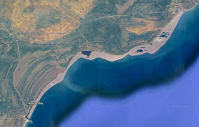 Figure 2. 1985 Google Earth image of the Gay mills stamp sand coastal deposit (gray unvegetated zone) extending from north of the village of Gay (upper right; mills site) to the Traverse River jetties at Traverse Bay village (lower left); a shoreline distance of over 5 miles. The large pond midway between Gay and the river jetties is the old Coal Dock site. The dominant subaerial deposit formed by sediment being carried from NE to SW and appears to terminate against the river’s north jetty. However, huge amounts of this waste material are deposited on the nearshore lake bottom, and the eroded suspended silts and clays are carried far offshore (e.g., greenish-blue zone along wider part of deposit and plume extending lakeward).