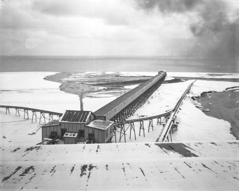 Figure 5. 1907 photo of the mills’ waste sediment disposal system showing sediment fans. In the center of the photo is the long concrete box-like flume structure that carried fine sediments (slimes) an boiler slag to the shore, and on each side conveyor belts that moved ‘sands’ into Lake Superior. Note the merging delta-like fans at the ends of the sluiceway and belts. As the emergent sand pile grew, the waste distribution system was extended lakeward, accreting a new shoreline up to over a half mile offshore. Remains of the flume and the north-side conveyor-system pilings still mark the Gay shoreline. (Photo courtesy of the J. Robert Van Pelt and John and Ruanne Opie Library, Michigan Technological University, Archives and Copper Country Historical Collections, MS042-050-999-U914C, http://www.mtu.edu/library/archives)