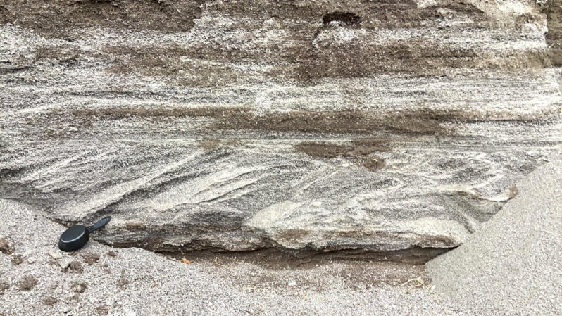 Figure 9. Close up of the wave-cut vertical scarp at the back of the beach showing bed forms in the original sluice deposit. Both horizontal beds with internal horizontal laminae (upper) and cross-bedded layers (lower) are common, reflecting the delta-like depositional conditions as the sluice sediments accumulated. Sampling cup for scale is 7.5 in (19.5 cm) in length.